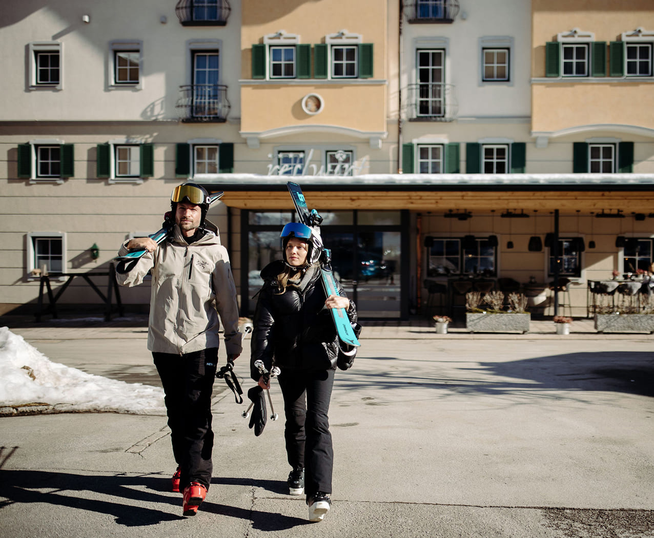 Paar geht im Sonnenschein mit geschulterten Skiern in Richtung Kamera.